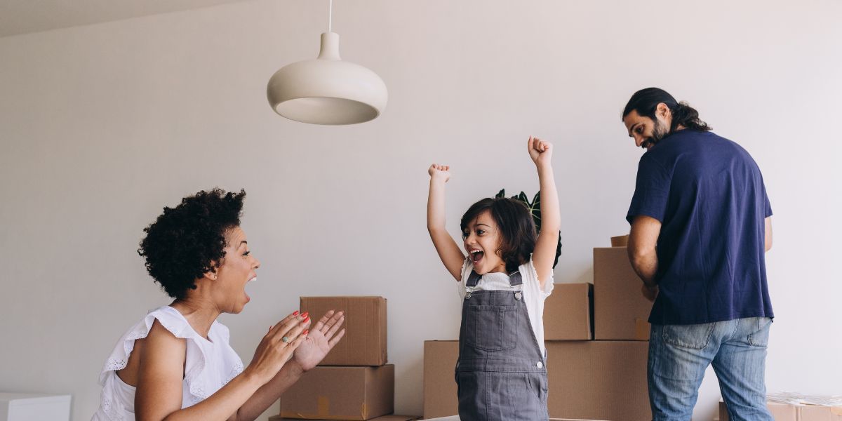 Excited child raising arms in joy while parents smile and unpack moving boxes in their new home.