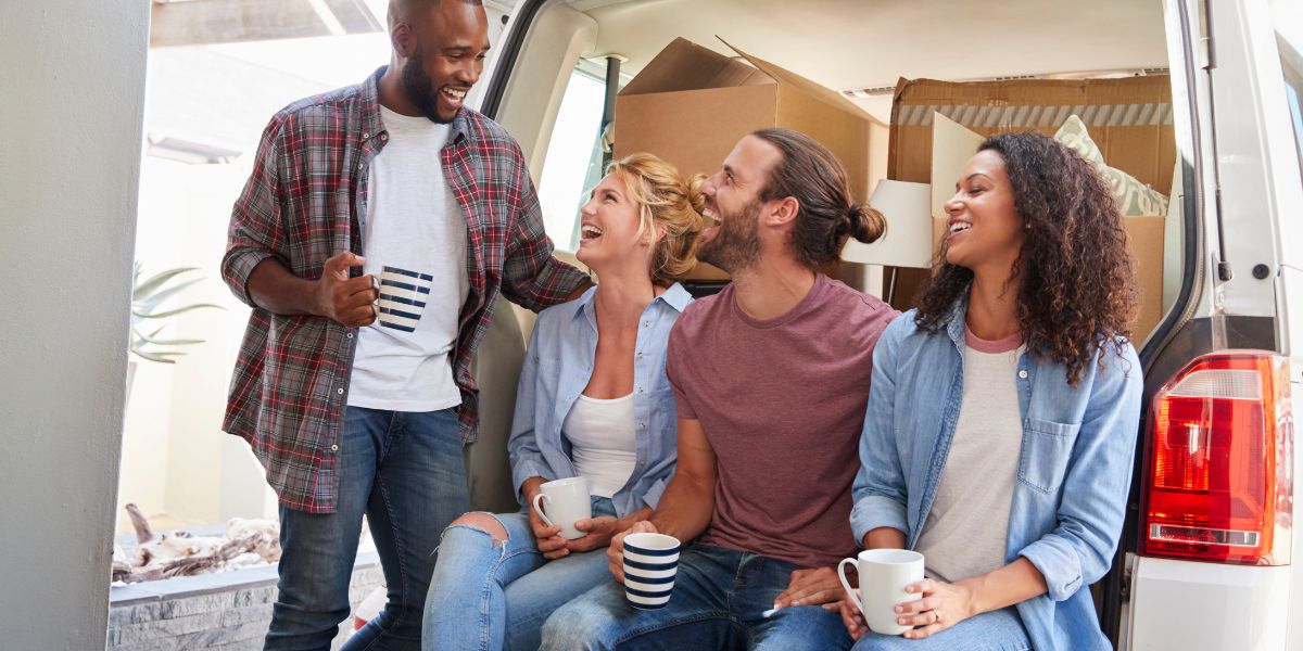 Group of friends sitting by a van, laughing and holding coffee mugs, surrounded by moving boxes.
