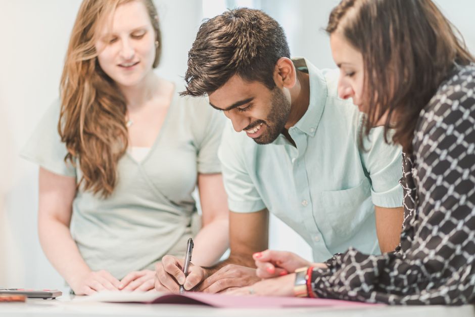 A happy couple sits at a modern wooden table, signing real estate contracts as their agent guides them through the paperwork.